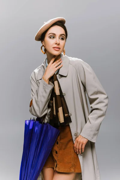 young woman in beret and trench coat posing with blue umbrella isolated on grey