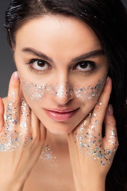 portrait of woman with natural makeup and glitter on cheeks and hands touching face while looking at camera isolated on grey clipart