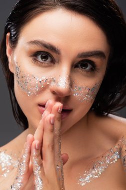 portrait of young woman with glitter on cheeks and praying hands looking at camera isolated on grey  clipart