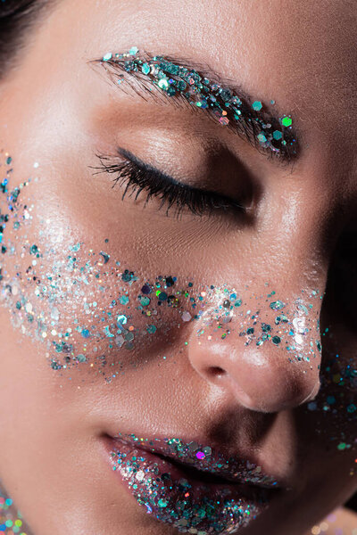 close up view of young woman with closed eye and sparking glitter on face isolated on black