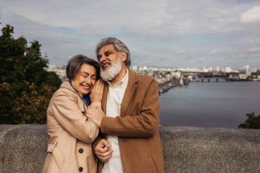 Son sınıf öğrencisi kadın paltolu kocasının omzuna yaslanıyor ve nehir kenarında gülüyor. 