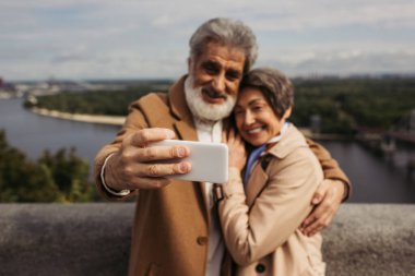 Mutlu bir son sınıf öğrencisi, mutlu bir eşle fotoğraf çekiyor. Trençkotlu, odaklanmış akıllı telefondan.