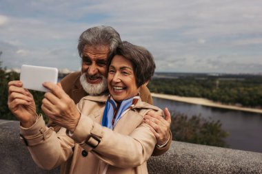 Trençkotlu neşeli yaşlı kadın ve sakallı kocasıyla selfie çekiyor. 