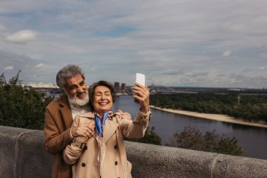 Bej renkli trençkotlu ve sakallı kocasıyla selfie çeken mutlu yaşlı kadın. 