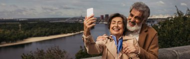 Bej renkli trençkotlu ve sakallı kocası ve pankartıyla selfie çeken mutlu yaşlı kadın.