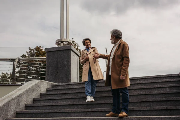 full length of stylish senior man in coat walking on stairs and holding hand of happy wife