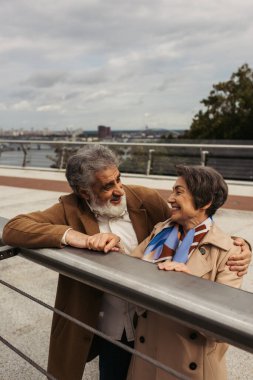 bearded senior man in coat hugging cheerful wife while standing near bridge guard rail  clipart