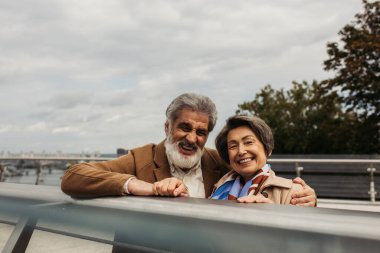 bearded senior man in coat hugging cheerful wife and standing near bridge guard rail  clipart