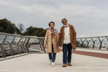 full length of happy senior couple in coats holding hands and walking on bridge near guard rail  clipart