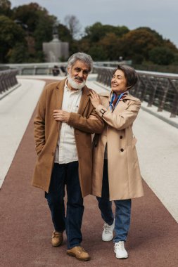 Köprüde yürüyen bej renkli sonbahar paltolu neşeli yaşlı çift.