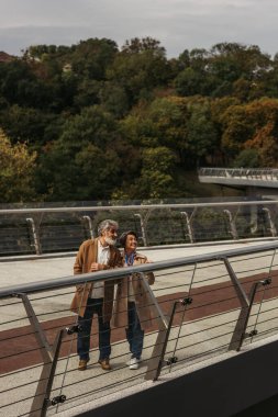 Uzun, mutlu yaşlı kadın ve sakallı adam köprü korkuluğunun yanında duruyor. 