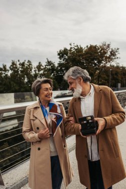 Köprüde gülümseyen karısının yanında eski model kamera tutan mutlu yaşlı adam. 