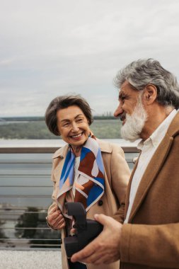 happy senior man holding vintage camera near curious wife smiling on bridge  clipart