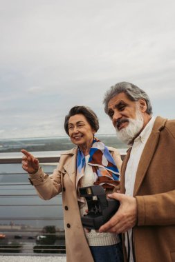 Eşinin yanında eski model kamera tutan mutlu yaşlı adam gülümsüyor ve köprüyü işaret ediyor. 