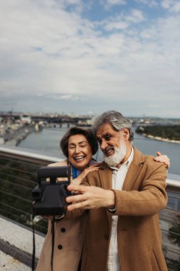 Mutlu son sınıf öğrencisi mutlu bir eş ve neşeli bir eş, arka planda bulanık bir şehir olan klasik bir kamera ile selfie çekiyor. 