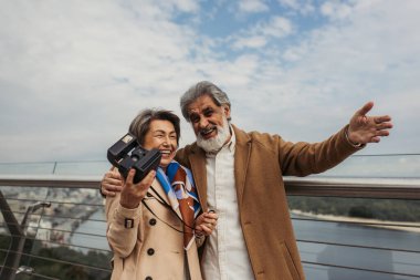 cheerful senior woman holding vintage camera while husband smiling and gesturing outside  clipart