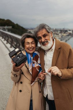 Trençkotlu neşeli yaşlı kadın elinde eski model kamerayla dışarıda mutlu bir koca tutuyor. 