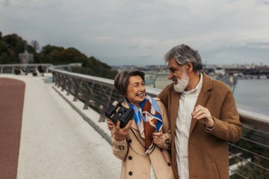 Trençkotlu neşeli yaşlı kadın elinde eski model kamera ve dışarıda mutlu kocaya bakıyor. 
