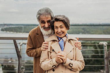 Sakallı neşeli adam dışarıda trençkotlu yaşlı karısına sarılıyor. 