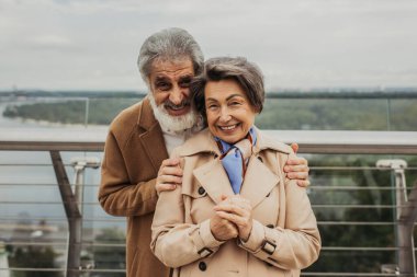 cheerful man with beard hugging senior wife in trench coat outdoors  clipart