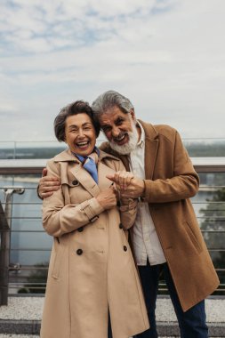 Sakallı neşeli bir adam dışarıda trençkotlu yaşlı karısına sarılıyor.