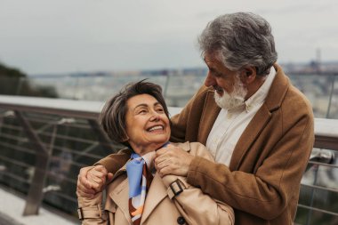 Mutlu kıdemli adam, bej ceketli pozitif karısına sarılıyor ve dışarıda gülümsüyor. 