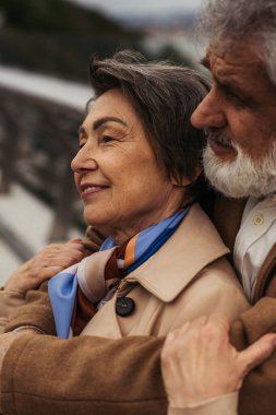 Sakallı yaşlı bir adamın portresi. Bej ceketli olumlu eşine sarılıyor ve dışarıda gülümsüyor. 