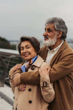 Sakallı yaşlı bir adamın bej renkli trençkotlu olumlu karısına sarıldığı ve dışarıda gülümsediği portresi. 