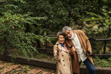 Sonbahar parkında ellerinde kağıt bardaklarla bej ceketli mutlu bir çift. 