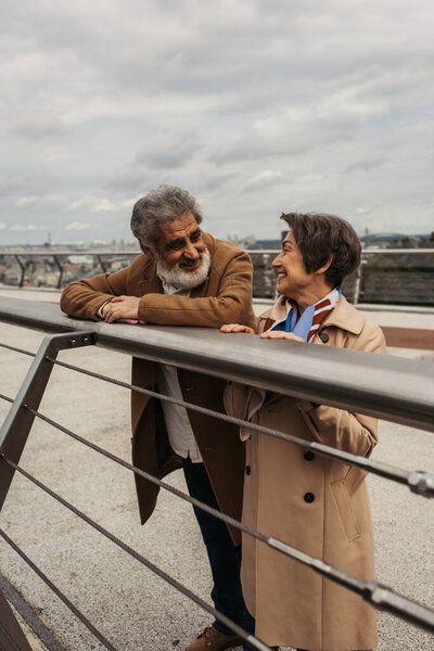 happy senior man in coat looking at pleased wife while leaning on guard rail of bridge 