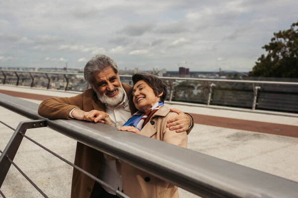 happy senior man in coat hugging cheerful wife while standing near bridge guard rail 