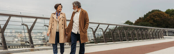 радостная пожилая пара в бежевых пальто, держась за руки и идя по мосту возле ограждения, баннер