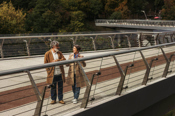 full length of happy senior woman and bearded smiling man standing near bridge guard rail 