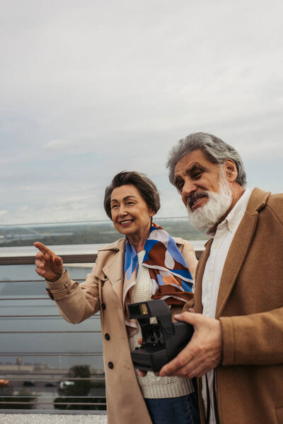 happy senior man holding vintage camera near wife smiling and pointing with finger on bridge 