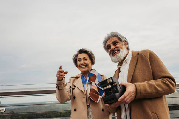 bearded and senior man holding vintage camera near wife smiling while pointing with finger on bridge 