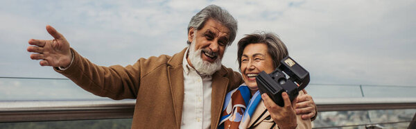 cheerful senior woman holding vintage camera while husband smiling and gesturing outside, banner