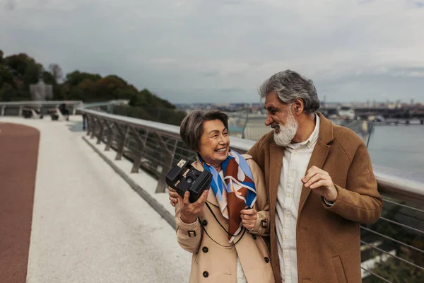 cheerful senior woman in trench coat holding vintage camera and looking at happy husband outside