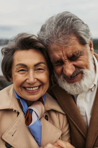 portrait of happy senior man and positive woman in beige coats smiling outside