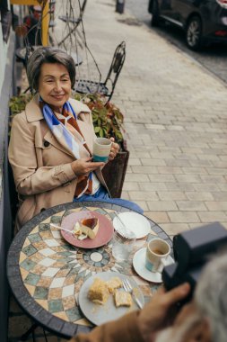 Yüksek açılı bir bakış açısıyla, yaşlı ve mutlu bir kadının eski bir kamerayla fotoğrafını çeken bir adam.