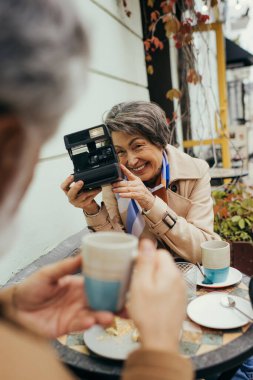 Elinde antika fotoğraf makinesi olan mutlu yaşlı bir kadın ve bulanık kocasının çay fincanıyla fotoğrafını çekiyor.