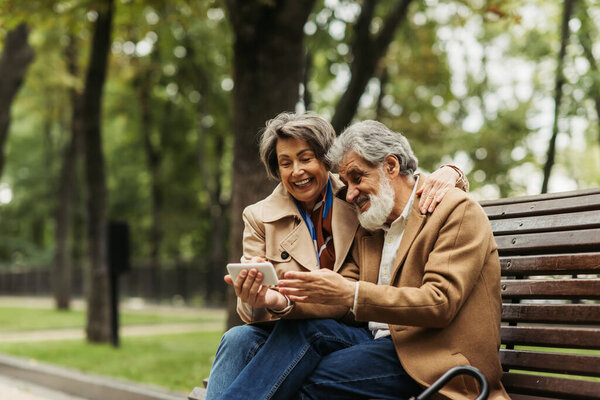 happy senior couple in coats sitting on bench and looking at smartphone 