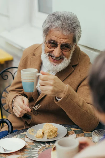 cheerful senior man in coat and eyeglasses holding cup of tea near cake and wife on terrace of cafe