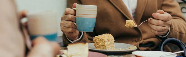 コートに身を包んだ年配の男性がお茶を飲みながらカフェのテラスで妻のそばでケーキを食べ — ストック写真