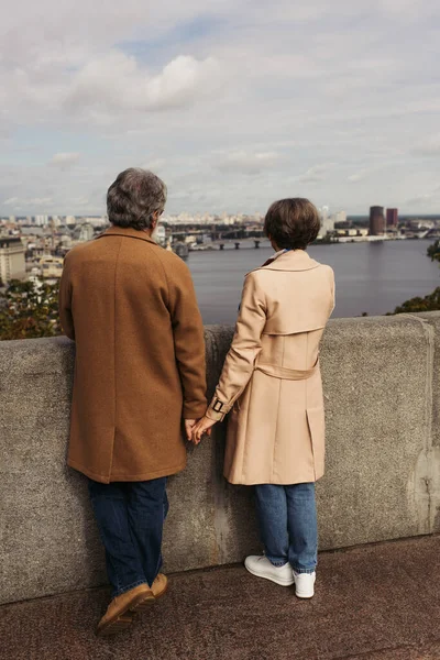 back view of retired couple in beige coats holding hands and standing on bridge near river with city view