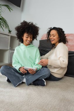 Neşeli Afrikalı Amerikalı anne halı üzerinde bacak bacak bacak üstüne atmış bir kızın ellerinde akıllı telefonu işaret ediyor. 