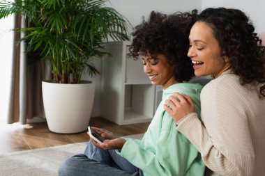 Mutlu Afro-Amerikan genç kız halının üzerinde oturuyor ve heyecanlı annesinin yanında akıllı telefon tutuyor.