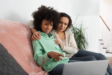 Kafası karışık Afro-Amerikan ergen kız laptopu kıvırcık annesinin yanına doğrultuyor.