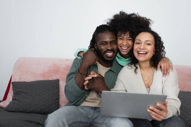 Mutlu Afro-Amerikan ailesi oturma odasındaki bilgisayarın yanındaki kameraya bakıp gülümsüyor. 