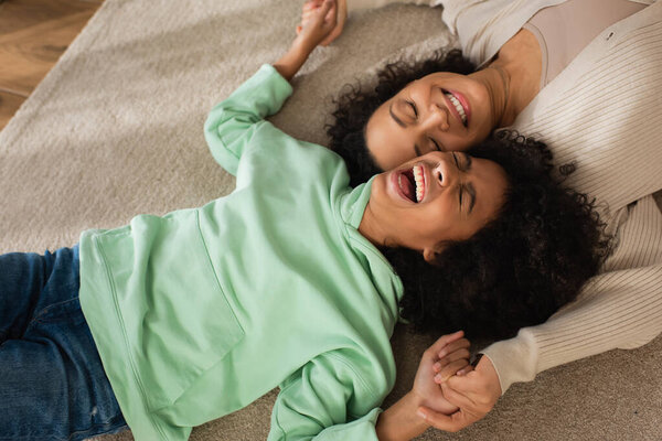 top view of happy african american girl laughing while lying on carpet with cheerful mother 