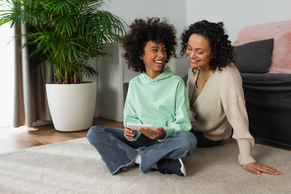 happy african american preteen girl sitting with crossed legs and holding smartphone near mother